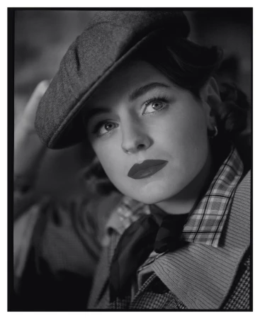 Monochromatic portrait of a woman wearing a newsboy cap, styled in a vintage fashion with red lipstick, captured in soft lighting.