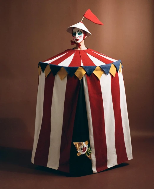 Person in a clown costume with a circus tent-like dress and pointed hat, standing against a brown backdrop for a theatrical presentation.