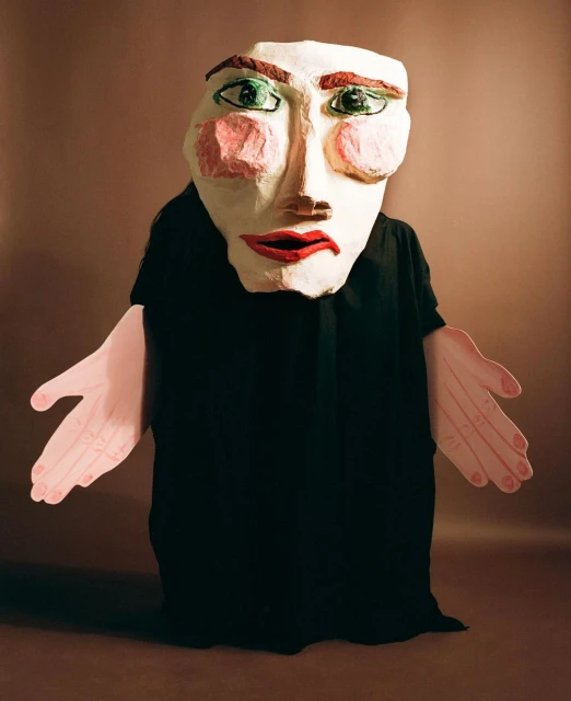 Person in a black robe wearing a large paper-mache mask with exaggerated features and pink paper hands against a brown background.