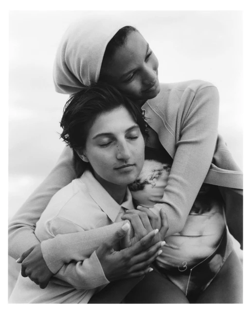 Black-and-white photograph of two women embracing; one with head covering looking down at the other with closed eyes in an outdoor setting.