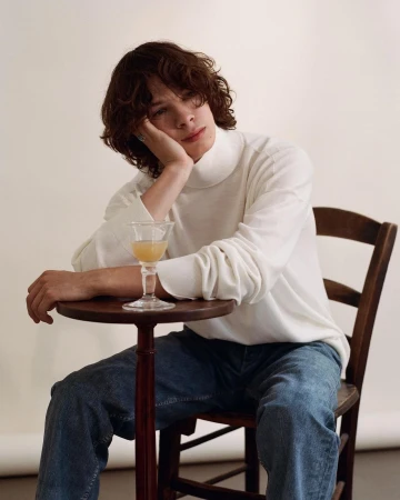 Young man with curly brown hair wearing a white turtleneck sweater and blue jeans sitting at a small wooden table.