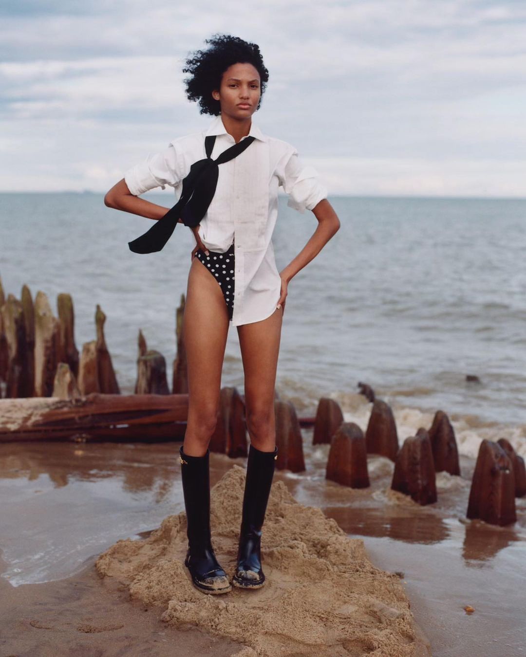 A young woman stands on a sandy beach near pilings wearing a white shirt, polka dot shorts, and tall black boots under a cloudy sky.
