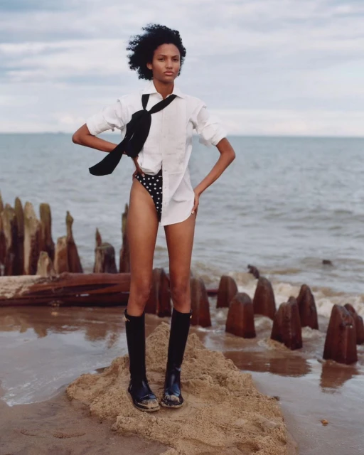 A young woman stands on a sandy beach near pilings wearing a white shirt, polka dot shorts, and tall black boots under a cloudy sky.
