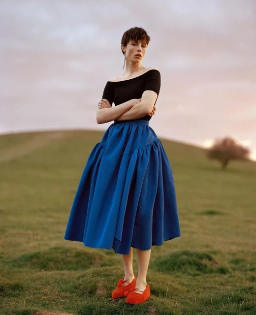 A woman in a black top and blue skirt stands in a field with green grass and a hill in the background, posing with her arms crossed.
