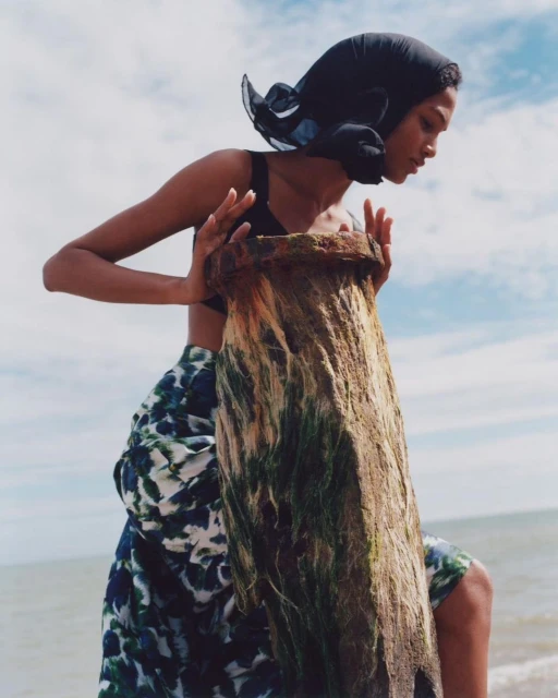 Fashion shot featuring an adult woman in a headscarf and swimsuit, posing with a weathered wooden post on a beach against a cloudy sky.