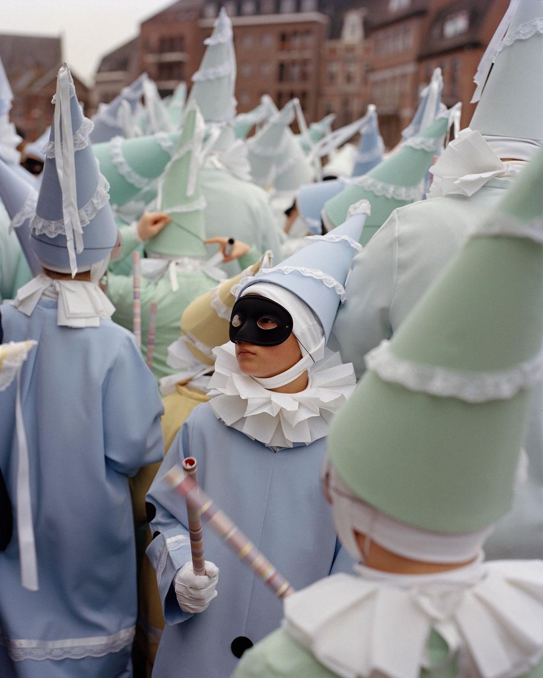 Parade of performers in costumes with pointed hats and black masks at an outdoor event in a European city setting on an overcast day.