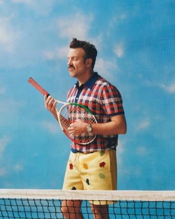 Jason Sudeikis holding a tennis racket wearing a plaid polo shirt and yellow shorts against a blue sky backdrop