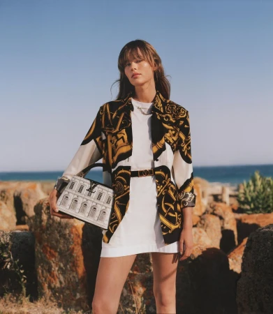 Model standing outdoors holding a structured white handbag wearing a printed patterned shirt and white dress against a coastal background.