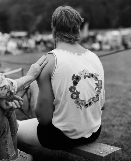 Black and white photo from behind showing a person in a tank top with a flower design sitting on a bench with another person's arm resting on their shoulder.