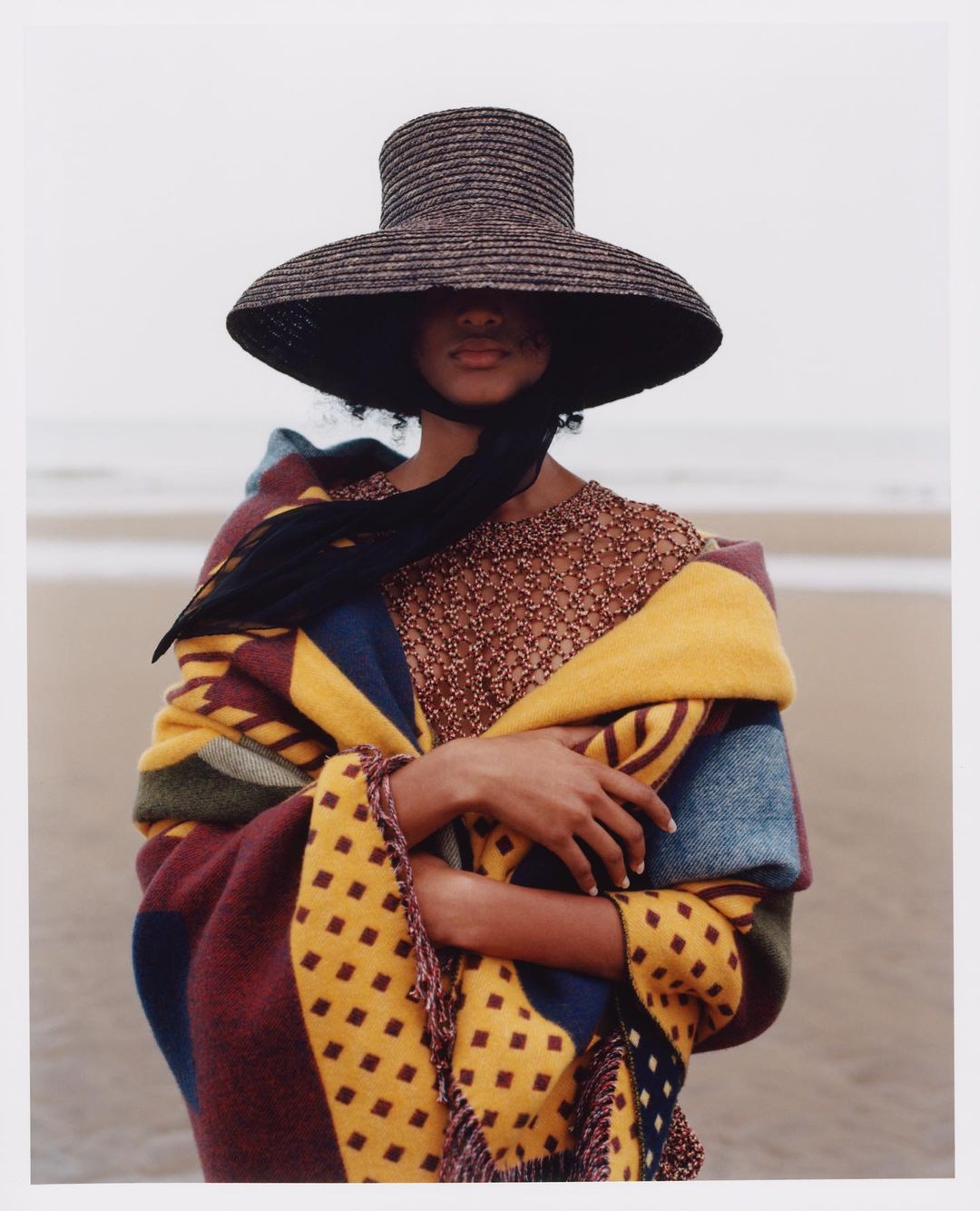 A woman wearing a straw hat and patterned shawl stands on a beach with waves visible in the background, showcasing a fashion or editorial aesthetic.