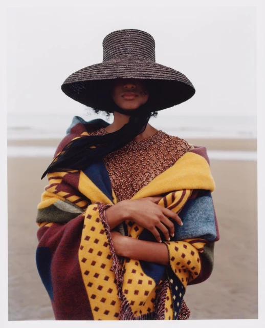 A woman wearing a straw hat and patterned shawl stands on a beach with waves visible in the background, showcasing a fashion or editorial aesthetic.