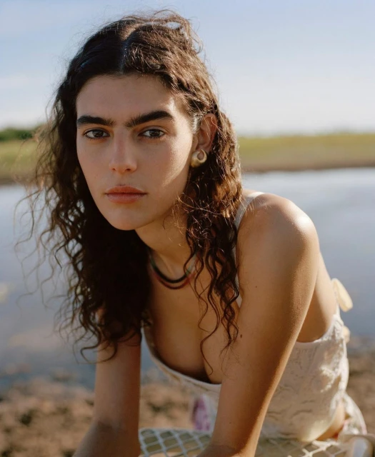 Close-up portrait of a woman with long, dark curly hair, wearing a floral-patterned top, posing outdoors near water under natural lighting.