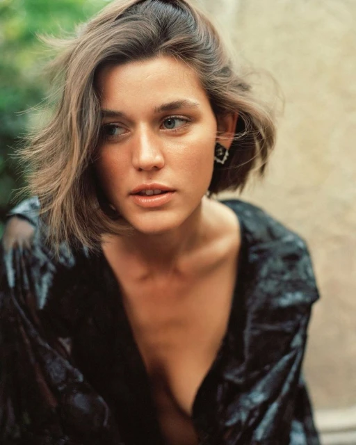 Portrait of a young adult woman with shoulder-length brown hair, wearing a black sheer top and earrings, in a soft outdoor setting.