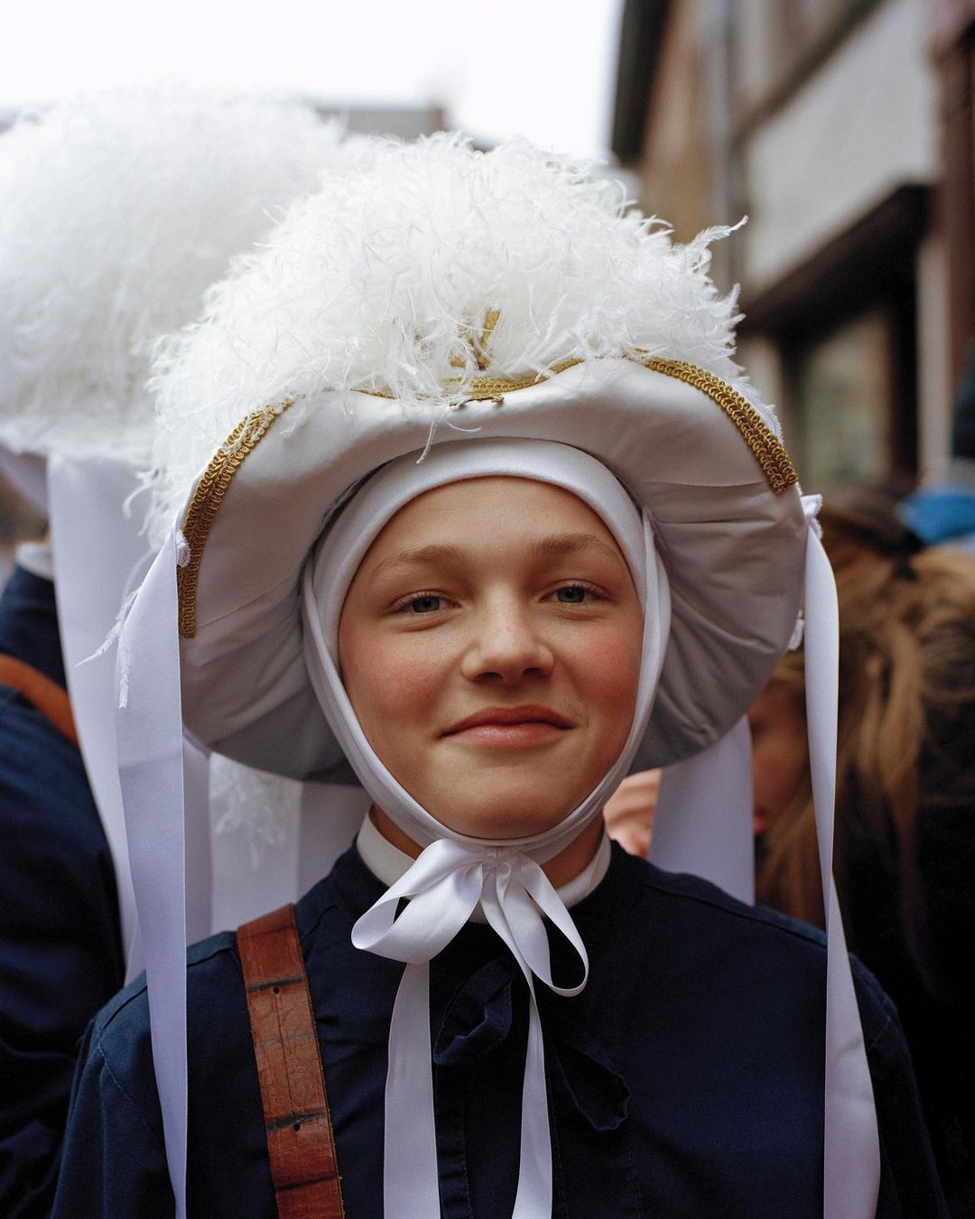 Close-up of a person in a traditional costume with a large feathered hat, white ribbons, and dark blue clothing, smiling at the camera.