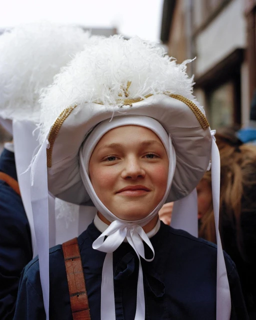 Close-up of a person in a traditional costume with a large feathered hat, white ribbons, and dark blue clothing, smiling at the camera.