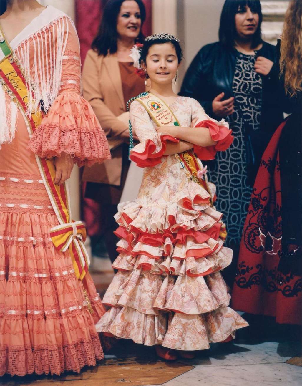 Young girl in traditional Spanish dress with ruffles and floral pattern, wearing sash and tiara, surrounded by other people in similar attire.