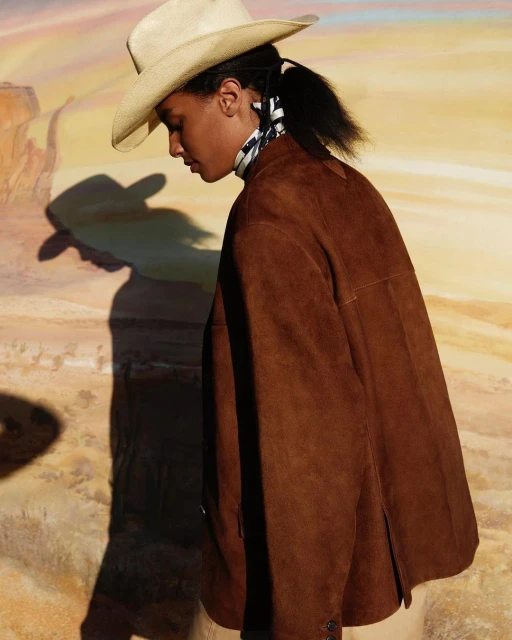 Woman in brown suede jacket and cowboy hat poses against a painted desert backdrop. Fashion shot with diffused lighting.