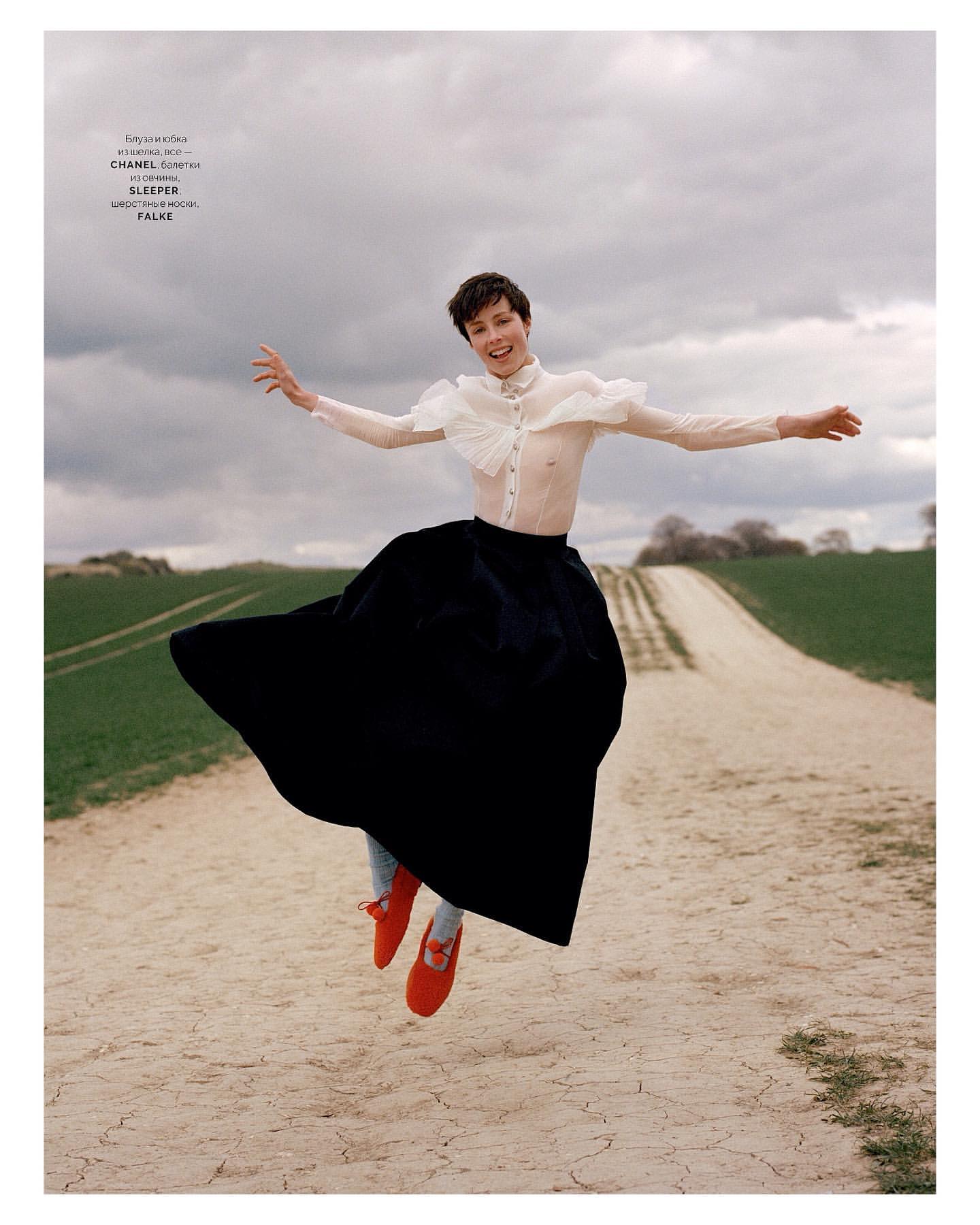 Young woman in a long black skirt and white ruffled blouse joyfully leaps on a dirt road, with a cloudy sky and green fields.