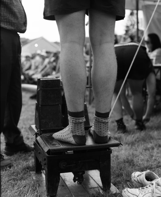 Person stands on small metal platform in a black and white outdoor shot, wearing socks, with a blurred background of people and tents.