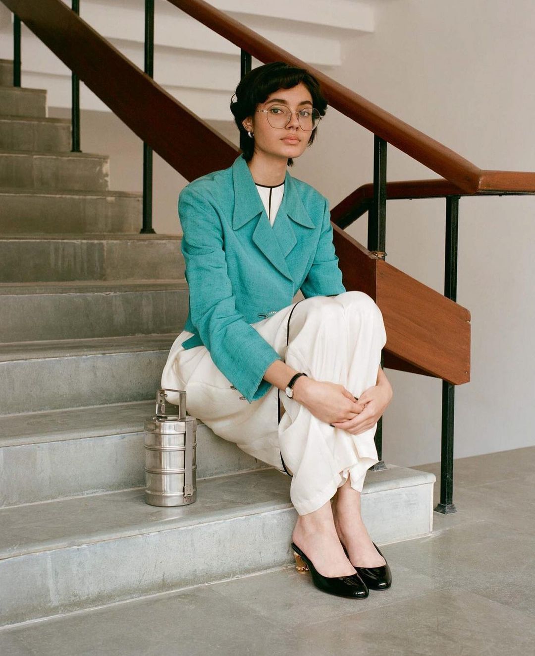 A young woman sits on a concrete staircase wearing a teal jacket and cream-colored pants with a lunch box and black heels.