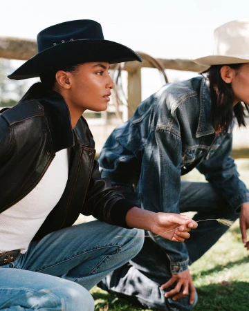 Two models wearing cowboy hats and denim crouching outdoors in a grassy landscape with a wooden fence.