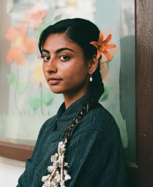 Portrait of a young woman with a flower in her hair, wearing a dark blue collared shirt, with a braided hairstyle and simple earring.