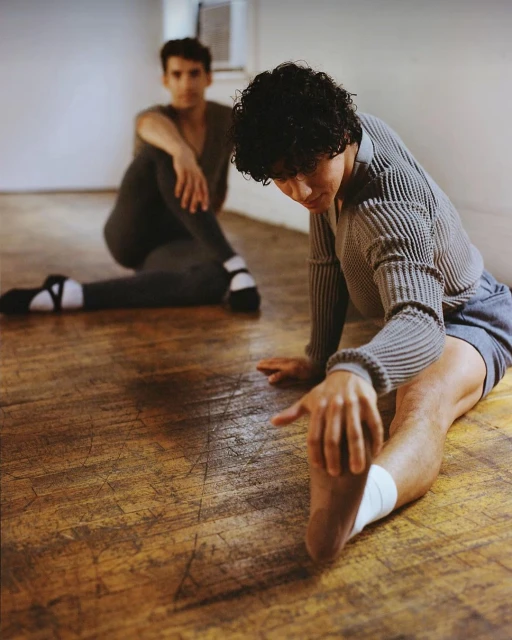 Two male dancers stretch in a studio, one with short curly hair in the foreground and another blurred in the background.