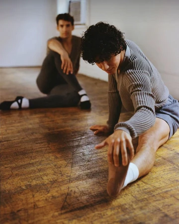 Two male dancers stretch in a studio, one with short curly hair in the foreground and another blurred in the background.