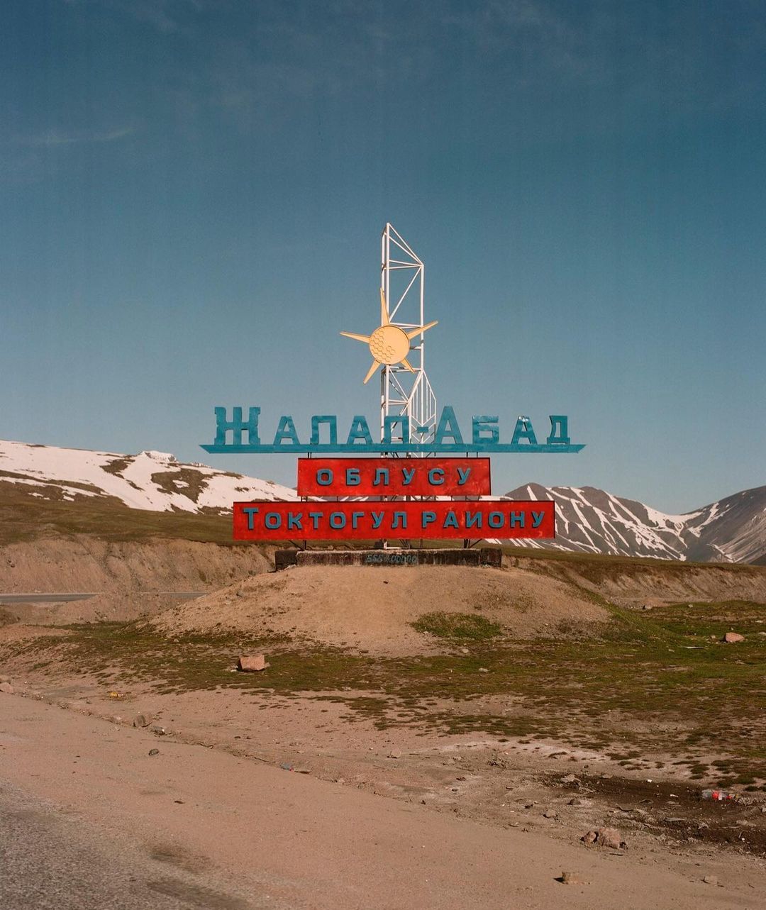 Sign in a hilly landscape with text in a foreign language, featuring a stylized sun and mountain backdrop under a clear sky.