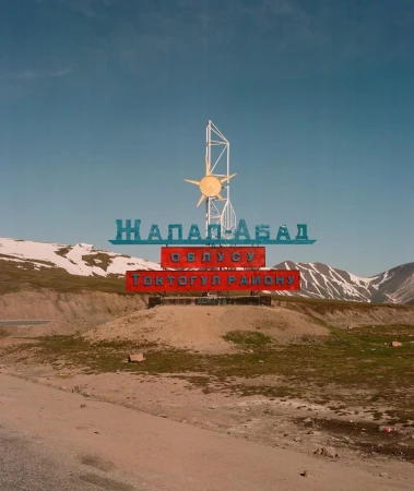 Sign in a hilly landscape with text in a foreign language, featuring a stylized sun and mountain backdrop under a clear sky. 