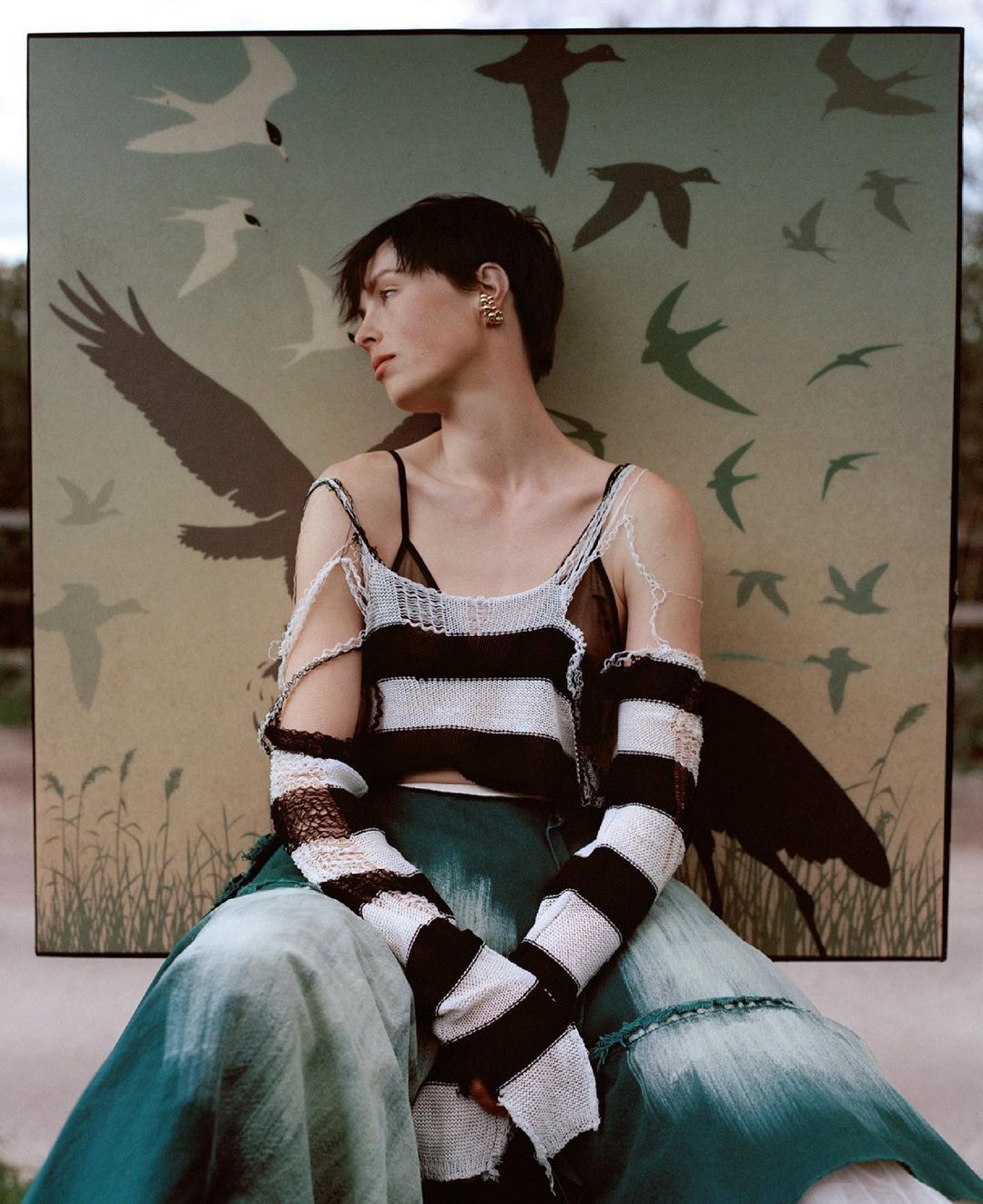 An adult woman poses in a black and white knit top and green tie-dye pants in front of a bird illustration backdrop in a close-up shot.