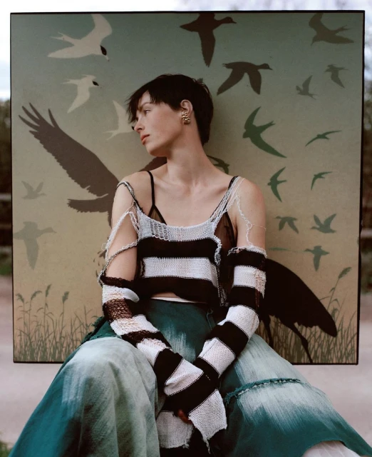 An adult woman poses in a black and white knit top and green tie-dye pants in front of a bird illustration backdrop in a close-up shot.
