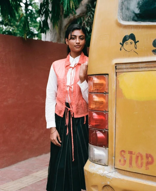 Young woman in a buttoned vest poses next to a yellow bus with illustrations; outdoor shot in daylight with a red wall backdrop.