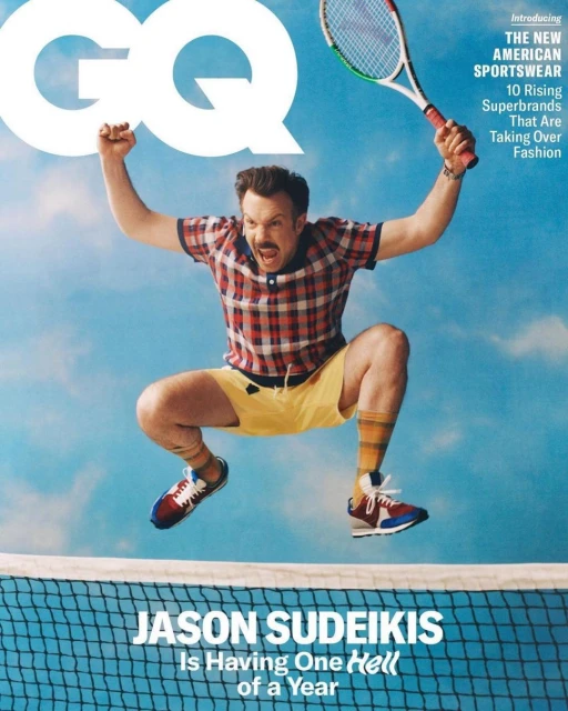 Magazine cover featuring Jason Sudeikis jumping in a tennis net with racket. He wears a red plaid shirt, yellow shorts, and tennis shoes.