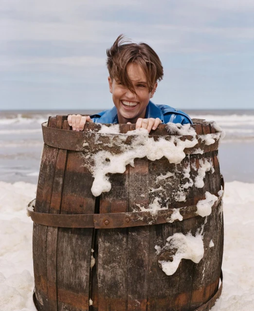 Smiling person in barrel covered with foam on beach near the ocean, wearing a blue shirt, with a cloudy blue sky.