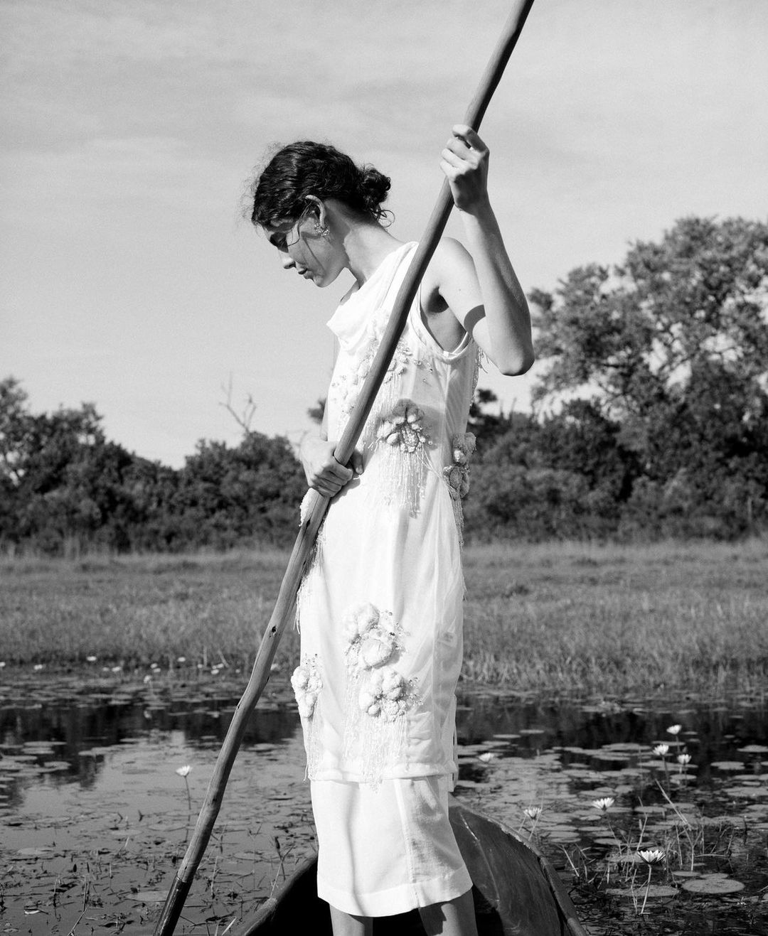 Black and white photograph of a woman in a white dress rowing a boat on a body of water with trees and sky in the background.