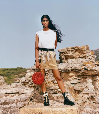 Model standing on stone ruins wearing a white sleeveless top, printed gold skirt, and black boots while holding a round red bag.