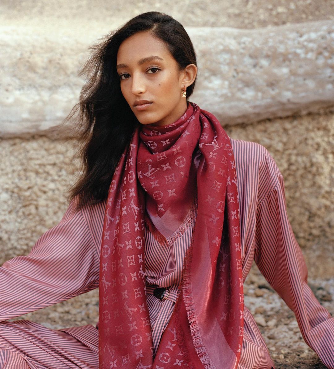 A woman wearing a patterned red scarf and striped outfit outdoors against a rocky background