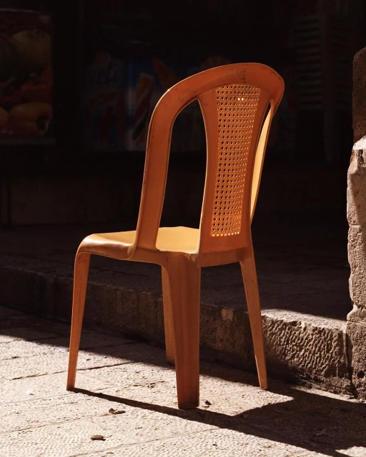 A close-up shot of a vacant orange plastic chair set outside on a stone surface with sunlight creating shadows and contrasts.