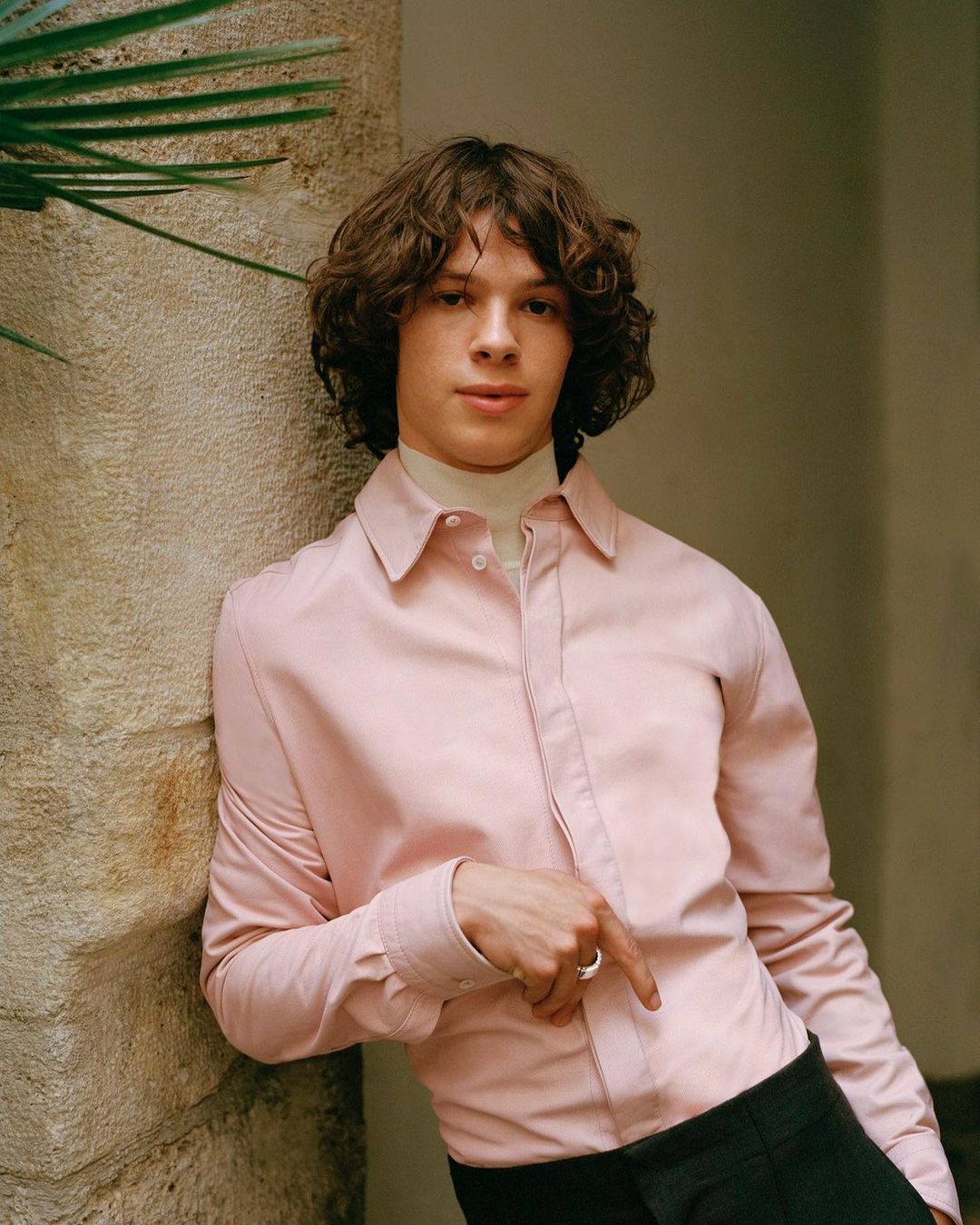 Young man with curly brown hair wearing a pale pink button-down shirt leaning against a stone wall