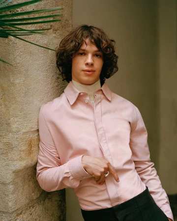Young man with curly brown hair wearing a pale pink button-down shirt leaning against a stone wall