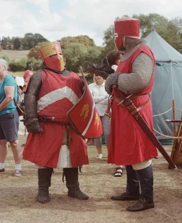 Two people in medieval knight costumes and chainmail stand in a grassy field near tents, with trees and people in the background under a cloudy sky.