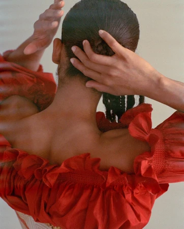 Rear view of a person with braided dark hair wearing a bright red ruffled top, adjusting their hair with both hands.