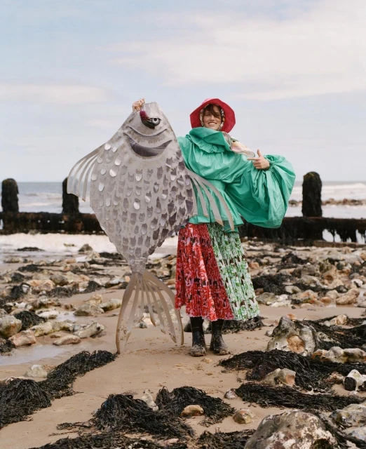A person on a beach with a large metal fish art piece, wearing a green jacket, a colorful skirt, and a red hat under a cloudy sky.