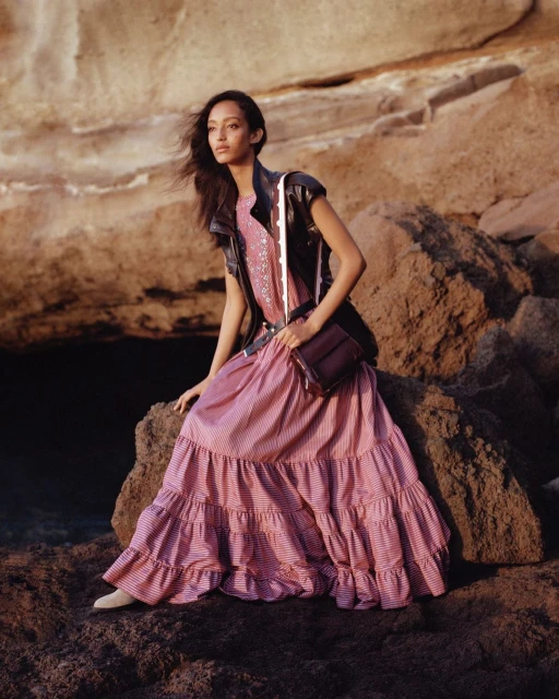 Woman in a pink striped dress and black vest sits on a rocky surface. Her dark brown purse sits on her lap against a tan rock background.