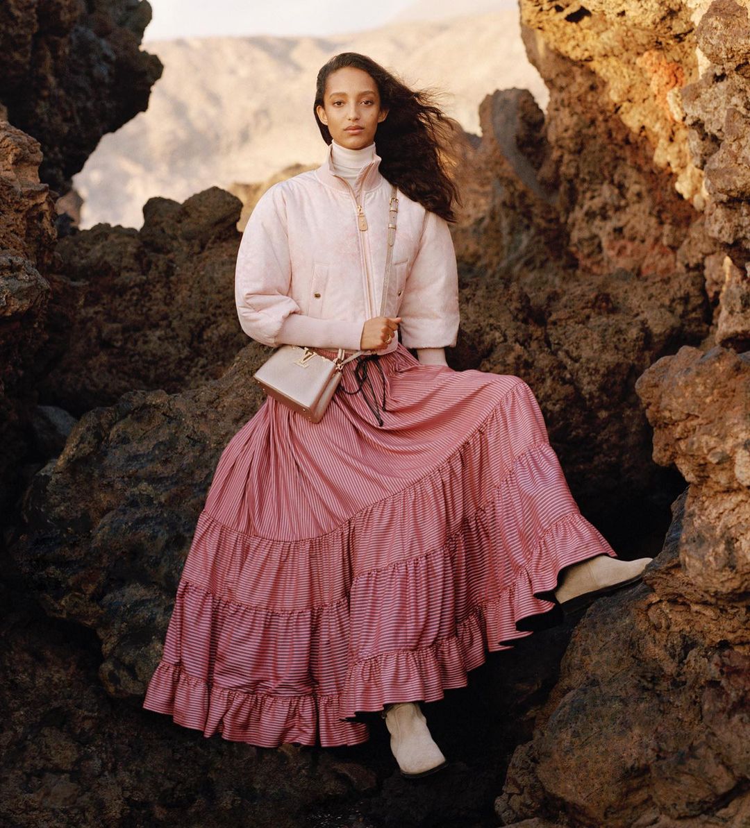 An adult female model poses outdoors among rocks, wearing a pink jacket, pink-striped skirt, and boots, accessorized with a purse.