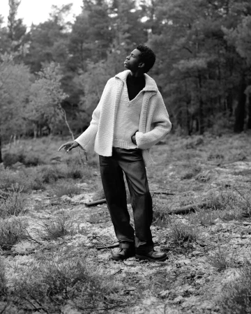 Black man wearing a textured knit cardigan and dark trousers standing outdoors among pine trees, photographed by Hill & Aubrey.