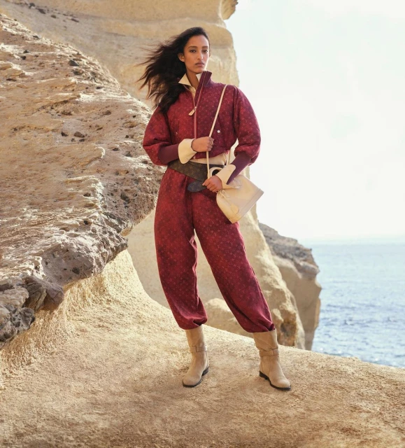An adult woman in a patterned maroon jumpsuit, boots, and shoulder bag poses on a rock formation near the ocean, with windblown dark hair.