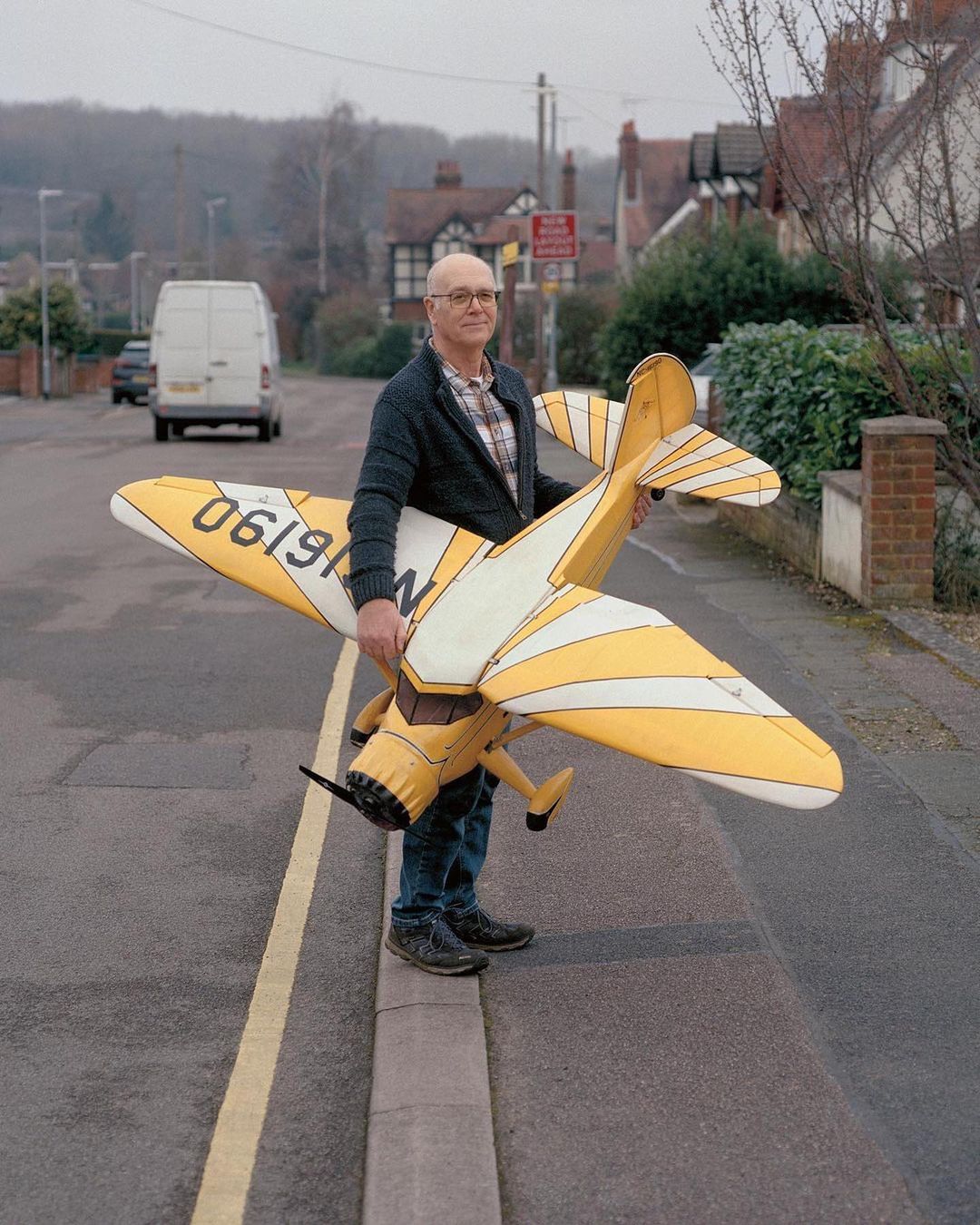 Man holds a yellow model airplane on a street with houses and a white van in the background, captured at eye-level.