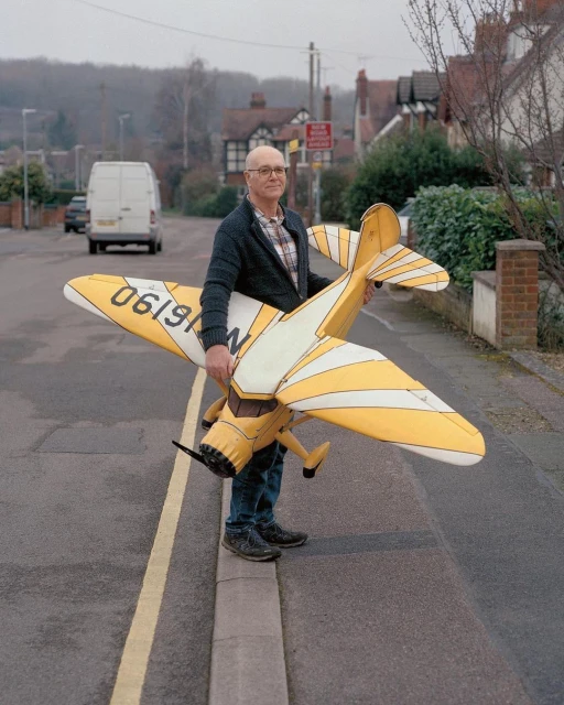 Man holds a yellow model airplane on a street with houses and a white van in the background, captured at eye-level.